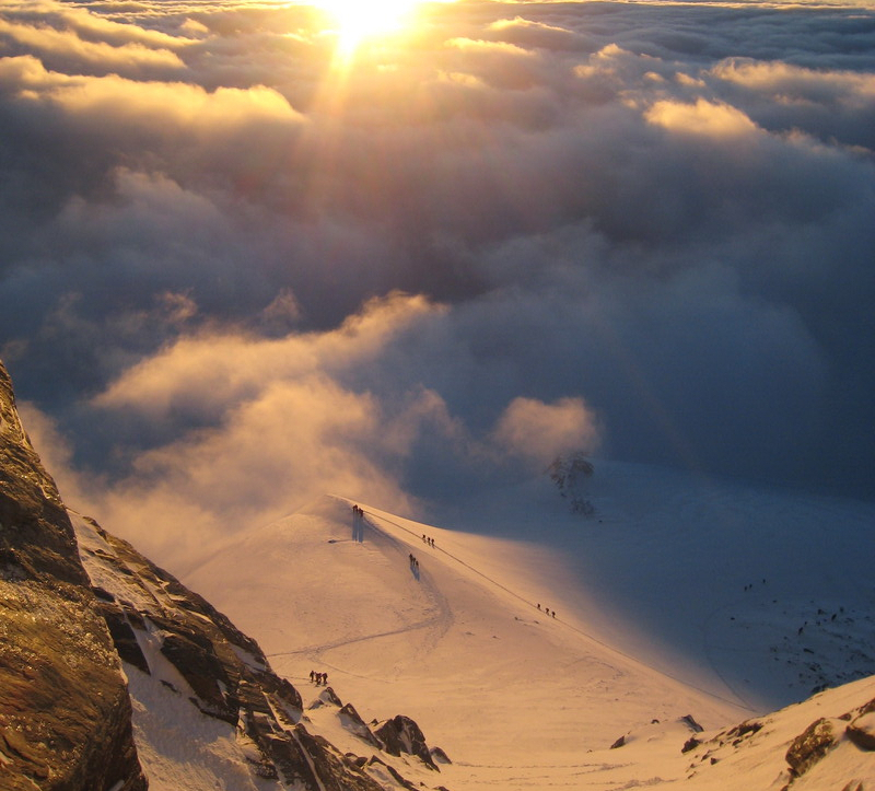 Sonnenaufgangsstimmung. Skibergsteiger:innen auf dem Weg zum Gipfel des Großglockners sind aus der Distanz zu erkennen. Es liegt Schnee. 