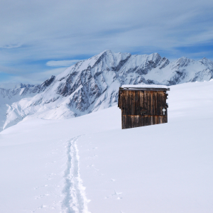 Eine Spur eines Skitourengehers im Vordergrund führt an einer kleinen Holzhütte links vorbei. Im Hintergrund Bergpanorama. 