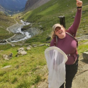 Eine junge Frau steht mit einem Insektenkäscher in den Bergen. Im Hintergrund ein Bach.