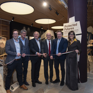 Vertreter der Nationalparks Dolomiti Belunese und Hohe Tauern schneiden bei der feierlichen Er&ouml;ffnung der Ausstellung &bdquo;Protected Futures&ldquo; ein rot-wei&szlig;-gr&uuml;nes Band in einem modern gestalteten Museumsraum mit Ausstellungselementen im Hintergrund.